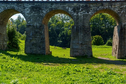A series of stone archways from an ancient structure, set against a lush green meadow with trees in the background. Scenic and peaceful historic landscape on a sunny day