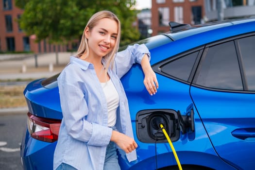 Young woman stands next to her electric car while charging it at a public station.