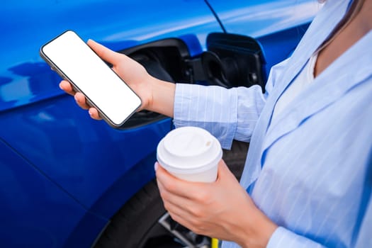 Woman charges their electric car, holding a coffee cup and checking their smartphone nearby