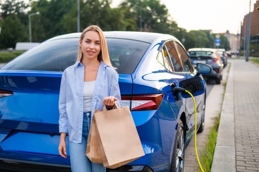 Woman smiles and holds a shopping bag while her electric car charges in a parking area.