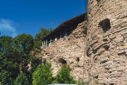 Close-up of an ancient fortress wall made of rugged stone, partially covered by greenery, with trees and a clear blue sky in the background. Historic and peaceful setting