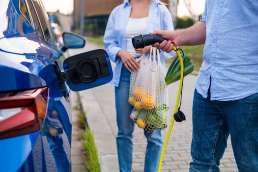Man connects an electric car to a charger while woman carrying a mesh bag of groceries