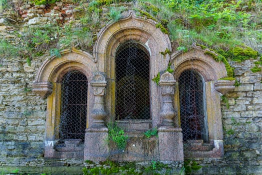 A close-up of an ancient stone facade featuring arched windows and overgrown with moss and vegetation, highlighting the weathered, historic architecture of a medieval fortress