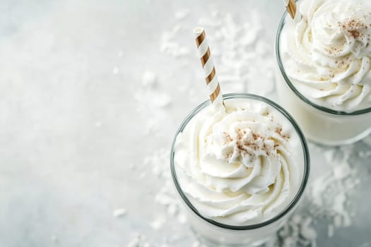 Two clear glasses filled with creamy beverages are topped with generous swirls of whipped cream, accented by striped straws. Shredded coconut decorates the surface.