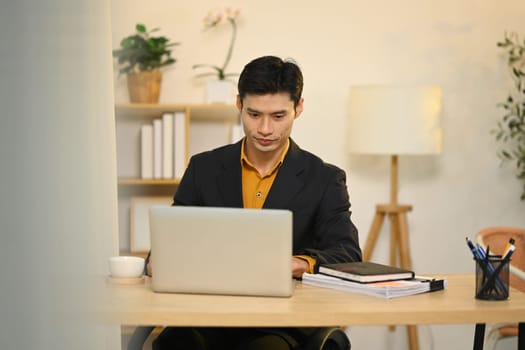 Concentrated asian businessman in black suit working on laptop at office.