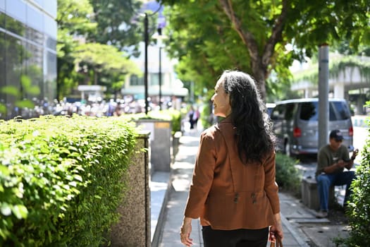 Back view of confident senior businesswoman strolling through urban greenery.