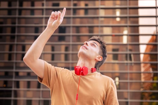 A young man wearing red headphones around his neck looks up and points with his finger, as if indicating something above. He stands in front of a modern building with a reflective glass facade.