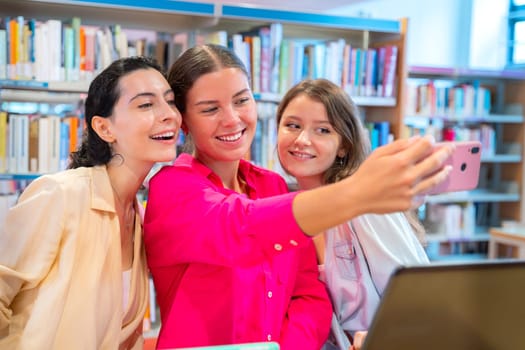 Three women smiling as they take a selfie together in a library setting. Surrounded by bookshelves, they capture a moment of joy,friendship, and study break, reflecting a positive learning environment