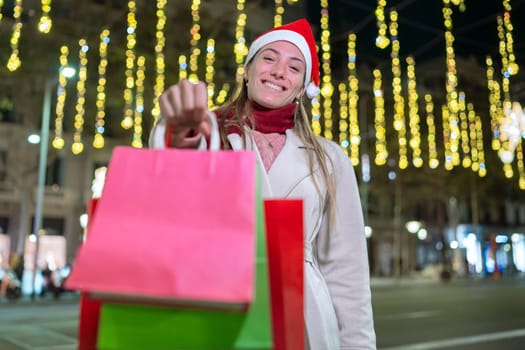 A cheerful woman wearing a Santa hat holds colorful shopping bags against a background of festive holiday lights. She is dressed warmly, enjoying a Christmas shopping night in the city.