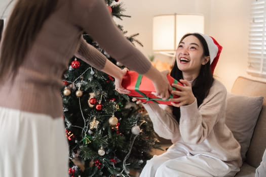 A young woman couple joyfully exchanges a surprise gift box by the Christmas tree, capturing the festive spirit in a cozy living room.
