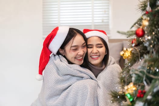 Happy young couple enjoying Christmas day, wrapped in blankets, wearing Santa hats, and smiling by a decorated tree.