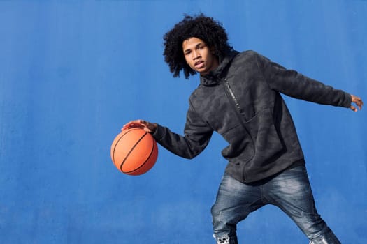 african american teen male in informal outfit playing basketball with ball near blue background looking at camera, concept of sport and young lifestyle, copy space for text