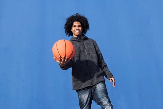 positive teen african american male with curly hair offering hand with basketball ball towards camera against blue background, concept of sport and young lifestyle, copy space for text