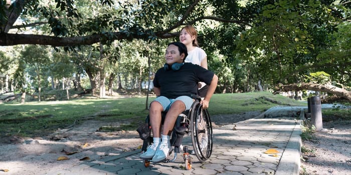A young woman helps a man in a wheelchair navigate a park path, enjoying the sunny day and natural surroundings.