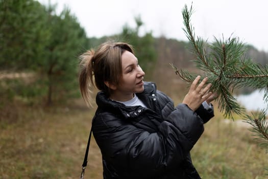 A woman in a black jacket inspects the branches of a pine tree while walking through a serene forest. The lake is visible in the background.
