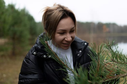 A woman is carefully collecting pine branches by the edge of a tranquil lake. The area is surrounded by lush trees under a gray, overcast sky, depicting autumn scenery.