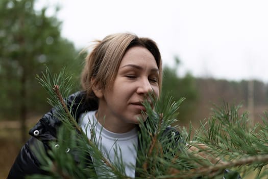 The woman leans in to smell the fresh pine branches, immersed in the serene atmosphere of the forest. Overcast skies create a calm backdrop for her nature walk.