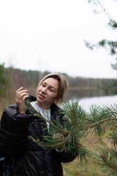 Near a tranquil lake, a woman inspects a pine branch while surrounded by lush greenery and soft autumn light, enjoying the natural environment.