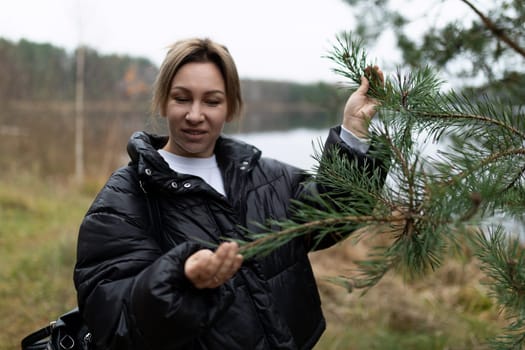 In a serene lakeside setting, a woman inspects a pine branch with curiosity, surrounded by the beauty of autumn foliage and tranquil waters.