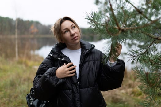 A woman stands by a calm lake, lightly touching the branches of a pine tree. Dressed in a cozy puffer jacket, she looks contemplatively towards the water on a cloudy day.