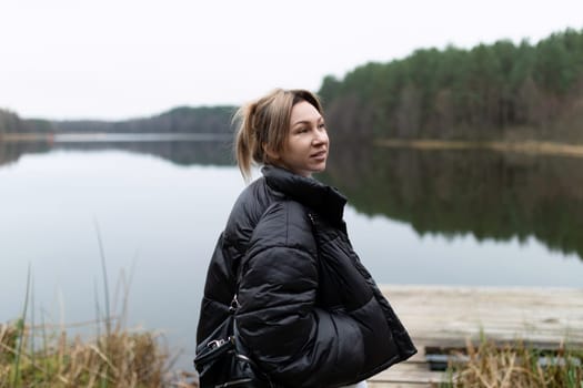 A young woman stands peacefully by a calm lake, wearing a warm jacket as she gazes thoughtfully at the serene surroundings of green trees and still water.