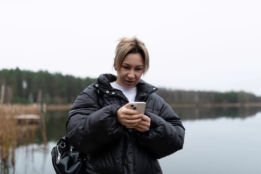 A woman with short blonde hair is standing by a lake. She is wearing a black puffer jacket and is looking at her phone. The weather is cloudy, and the lake is calm.