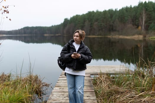 A woman in a black jacket and blue jeans walks along a wooden dock over a calm, reflective lake. She holds a smartphone in her hands and is looking towards the right side of the frame.