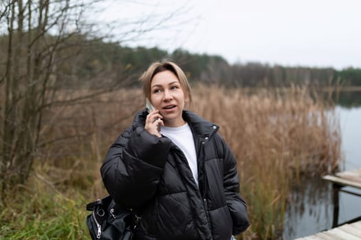 A woman is standing by a lake, wearing a black puffer jacket and holding a phone to her ear. She has short blonde hair and is looking away from the camera.