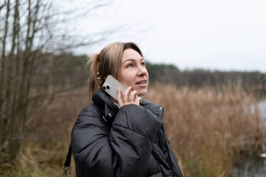 A woman with blonde hair, wearing a black puffer jacket and a strap across her body, is standing in a field with tall grasses.