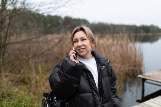 A woman wearing a black puffer jacket and a white shirt stands near a lake. She holds a phone to her ear with her right hand. The lake is behind her, and tall grass grows near the water.