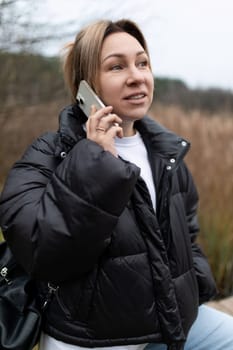 A woman with blonde hair, wearing a black puffer jacket and a white shirt underneath, holds a phone to her ear while talking.