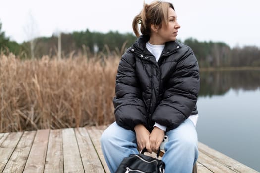A woman wearing a black puffer jacket and blue jeans sits on a wooden dock. She is looking out at a lake, her hands resting on a black bag in her lap. There is a field of tall grass behind her.