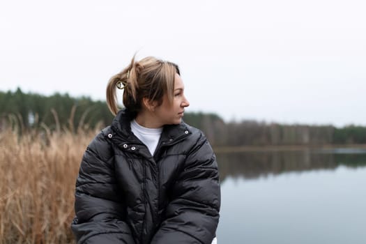A woman with light brown hair pulled back in a ponytail is wearing a black puffer jacket and looking to the right, over a calm lake. There are trees and tall grass in the background.