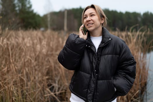 A young woman laughs while conversing on her phone near a calm lake, framed by tall grasses and trees, capturing a cozy autumn day outdoors.