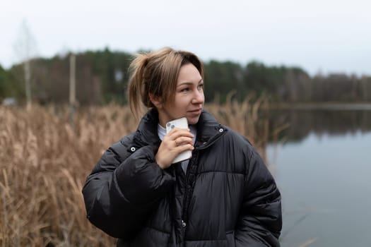 The young woman stands thoughtfully near the lake's edge, wrapped in a warm black puffer jacket, casually holding her phone as she gazes across the water surrounded by reeds.