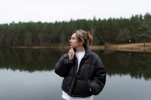 A young woman thoughtfully gazes over a serene lake reflecting the autumn sky, with dense pine trees in the background on a tranquil, cloudy day.