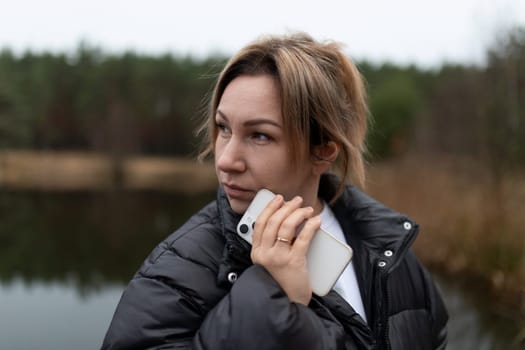 A woman wearing a black jacket is positioned near a calm lake, engaged in a phone conversation as gray clouds drift overhead, creating a tranquil atmosphere.