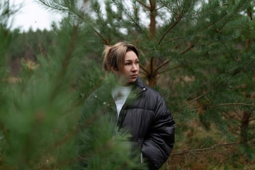 A woman in a black jacket observes her surroundings in a serene forest setting, surrounded by lush green trees, capturing a moment of peace and introspection.