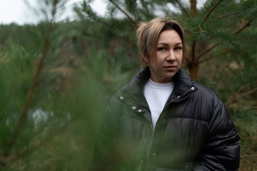 Amidst dense greenery, a woman dressed in a black jacket gazes thoughtfully, embracing the serene atmosphere of the forest on a cool day.