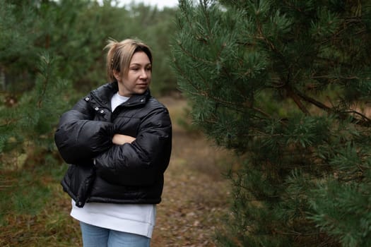 A woman wearing a cozy black puffer jacket stands with her arms crossed, gazing thoughtfully as she enjoys a tranquil moment among tall pine trees in the forest.