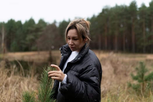 In a serene forest, a woman gently inspects the needles of a pine tree, surrounded by tall trees and grasses on a cool, cloudy day, reflecting on nature's beauty.