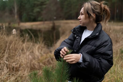 A woman dressed in a black puffer jacket is carefully handling small branches of a tree near a tranquil lake surrounded by autumn foliage. The setting is peaceful and serene.