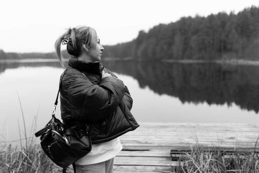 A young woman looks thoughtfully at the calm lake while standing on the wooden dock. The landscape features dense trees, creating a serene atmosphere.