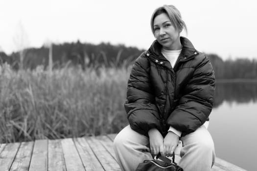 The woman sits on a wooden dock beside a serene lake, surrounded by tall grass and trees, wearing a warm puffy jacket and relaxed pants, lost in thought.