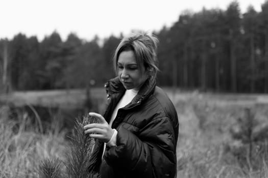 A woman in a puffy jacket walks through a forest, gently touching the branches of a pine tree, reflecting on the tranquil beauty of nature.