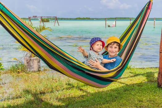 Two boys, a baby and a teenager lying in a hammock against the backdrop of turquoise waters of Bacalar Lake, Mexico. Peaceful tropical travel destination concept.