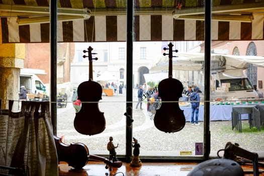 Cremona, Lombardy, Italy - November 6th 2024 Two violins are displayed in a luthier's shop window, with an outdoor market visible in the background
