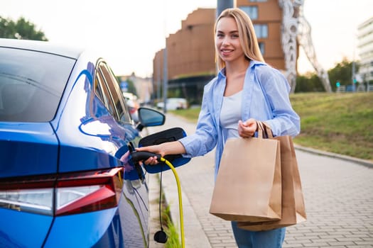 Portrait of blonde woman plugs in her electric vehicle to charge while holding multiple shopping bags.
