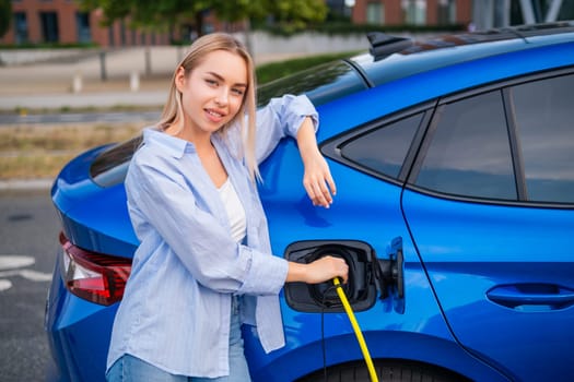 Portrait of blonde woman in casual clothes charging her electric car at a public parking station