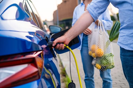 Man plugs in an electric car for charging while woman holds a grocery bag in the parking lot.